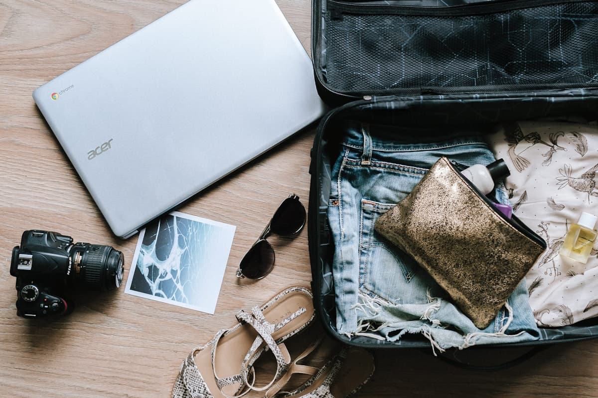 An open suitcase on a wooden floor beside a camera, sunglasses and a laptop, with a toiletry bag inside